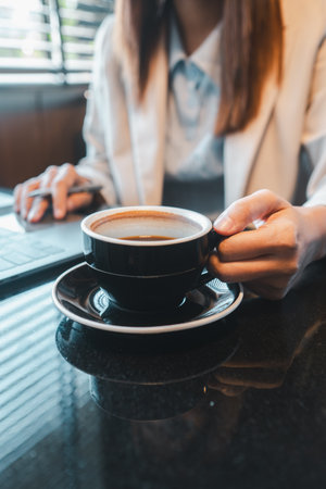 Professional woman holding coffee cup while using laptop, symbolizing productivity and relaxation in a modern workspace.の写真素材
