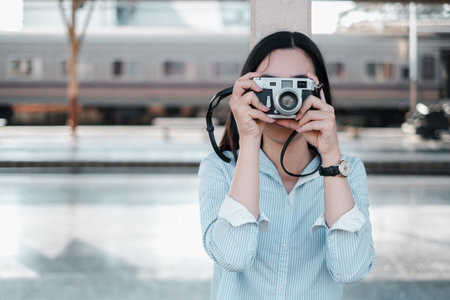 A woman in a blue shirt holds a vintage camera at a train station, ready to capture memories.