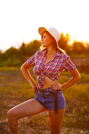 American young woman in plaid shirt and denim shorts, hat at sunset looks into distanceの写真素材