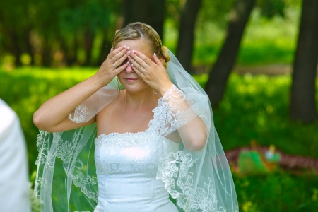 bride woman has closed eyes with his hands for a surprise, mystery in a romantic atmosphere in forest on a green background on your wedding dayの写真素材