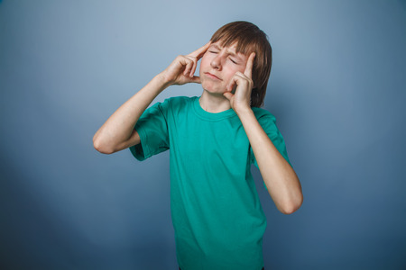 boy teenager European appearance brown hair in a turquoise t-shirt pressed his fingers to his temple on a gray background, thinking, thoughtの写真素材