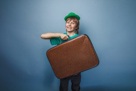 European appearance teenager boy in a shirt in green hat holds an old suitcase in hand on gray background, adventureの写真素材