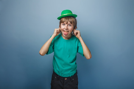 European appearance teenager boy in T-shirt with green hat stretches out his tongue and ears on gray background, foolery, teasingの写真素材