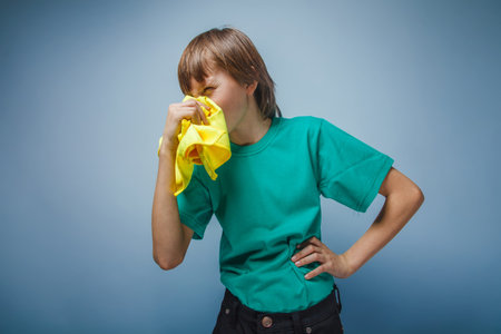 boy teenager European appearance brown hair blowing his nose in a T-shirt in yellow towel on a gray background, flu, cold, diseaseの写真素材