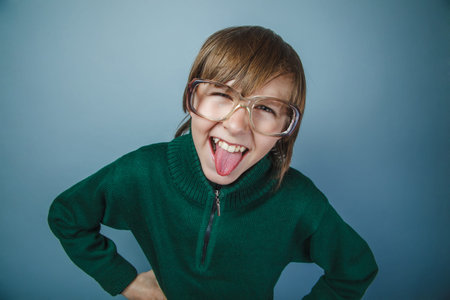 European-looking boy of ten years in glasses showing tongue on a gray background