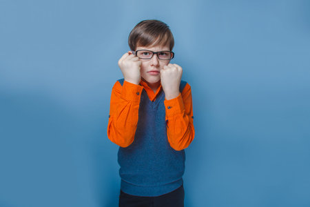 European-looking boy of ten  years  in glasses frowning, unhappy shows fists on a blue background