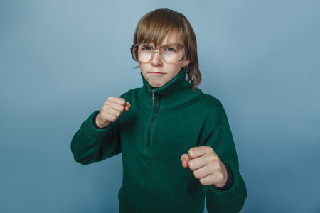 European- looking boy  of ten  years  in glasses frowning, unhappy shows fists on a blue background