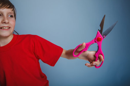 boy teenager European appearance in a red shirt holding a brown pink scissors on a gray background,...