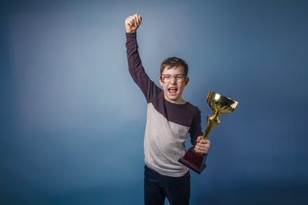 boy teenager European appearance in sunglasses in brown hair jacket holding a golden cup raised his hand up on a gray background shouts, joy, victoryの写真素材
