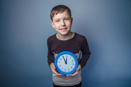 boy teenager European appearance in brown sweater holding a blue clock on a gray background, smileの写真素材