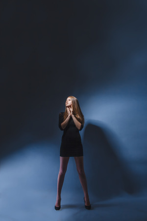 European appearance blonde girl with glasses in black dress legs wide apart leaned hands to her face on a dark gray backgroundの写真素材