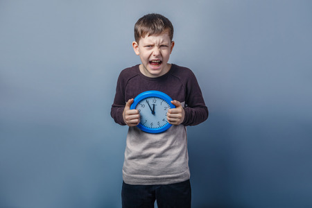 European-looking boy of ten years holding a wall clock opened his mouth  on a gray backgroundの写真素材