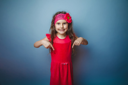 Girl European appearance haired child of seven in red bright dress shows thumb down on the gray backgroundの写真素材