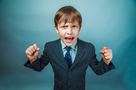 Teen boy businessman in a suit swearing angry shouts on a blue background photo studioの写真素材