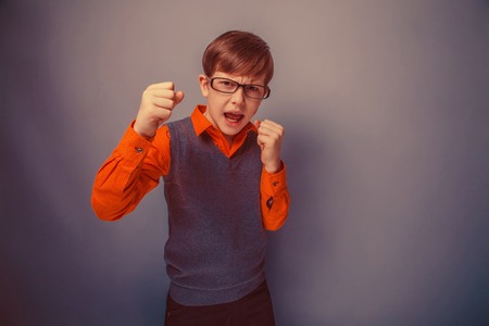 European-looking boy of ten years in glasses, anger, opened his mouth, his fists on a gray background retroの写真素材