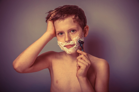 Teen boy shirtless European appearance in brown hair foam on face shaving on a gray background, a child growing up retroの写真素材