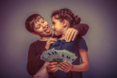 teen girl about seven years old holding a money boy teen hugging her shows tongue on a gray background, notes, fun, tease retroの写真素材