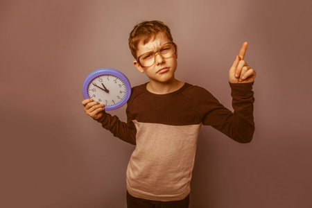 European-looking boy of ten years in glasses holding a wall clock on a gray background retroの写真素材
