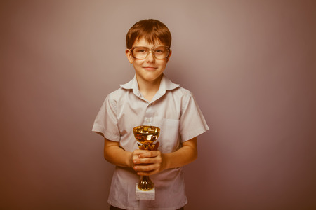 Teen boy brown hair European appearance in sunglasses holding her hands a golden cup on a gray background, happiness, reward retroの写真素材