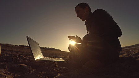 businessman man freelancer working behind sitting on beach freelancing laptop silhouette in the sunの写真素材