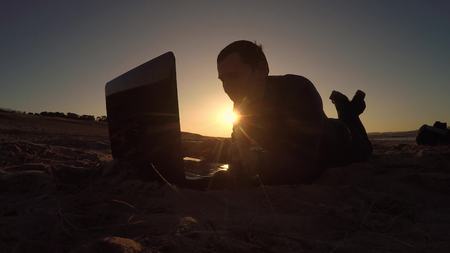 man laptop. businessman freelancer man laptop working behind sitting on beach freelancing silhouette in the sunの写真素材