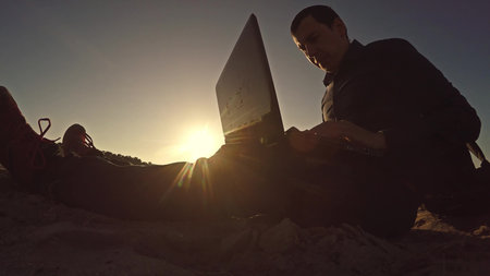 man businessman freelancer working behind laptop sitting on beach silhouette in the sun freelancingの写真素材