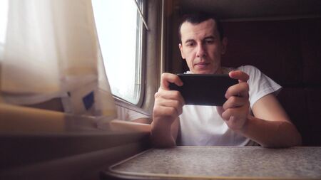 man businessman with smartphone travelling by train. man traveler is typing internet web message in train on the railway en route sits by the window of carriage. railroad travel concept. slow motion lifestyle video. interior inside trainの写真素材