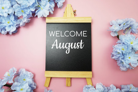 Welcome August text on wooden blackboard and flower decoration on pink background