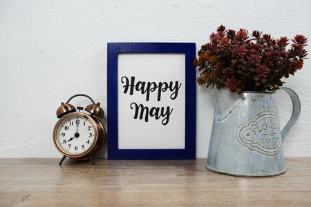Happy May typography text with alarm clock and artificial plant decoration on wooden table and white wall backgroundの写真素材