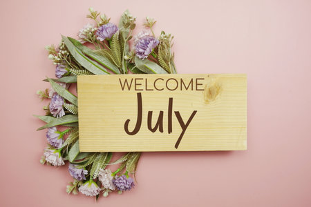 Welcome July text on wooden board with flowers frame on pink backgroundの写真素材
