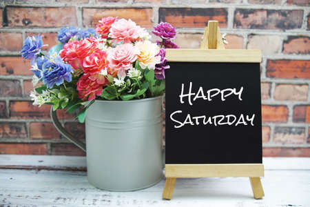 Happy Saturday text on black wooden easel and vase with flowers bouquet on brick wall backgroundの写真素材