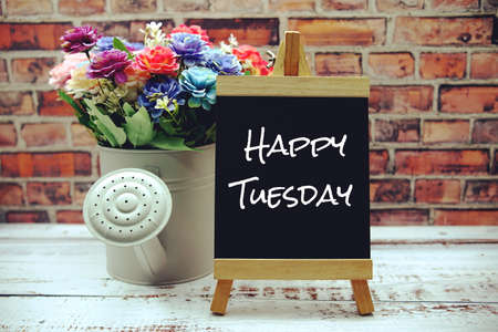 Happy Tuesday text on black wooden easel and vase with flowers bouquet on brick wall backgroundの写真素材