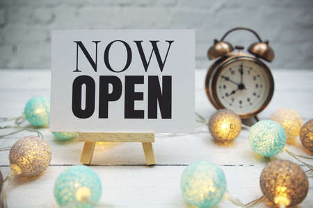 Now Open text and alarm clock on white brick wall and wooden backgroundの写真素材