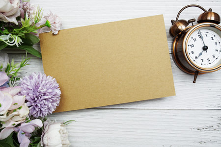 Empty paper card with flower and alarm clock flat lay on wooden background