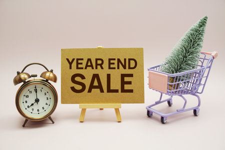 Year end sale text message with wooden easel and alarm clcok, christmas tree on trolley shopping cart on pink backgroundの写真素材