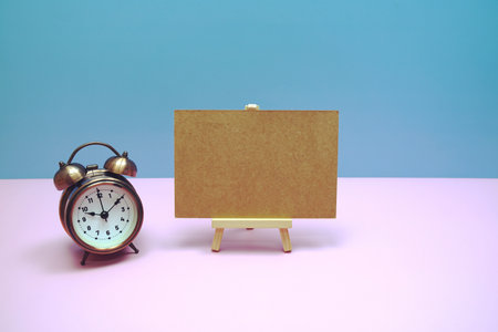 Empty card mockup space for text message with wooden easel and vintage alarm clock on pink background, business and education backgroundの写真素材