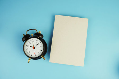 Black alarm clock with empty paper card top view on blue background