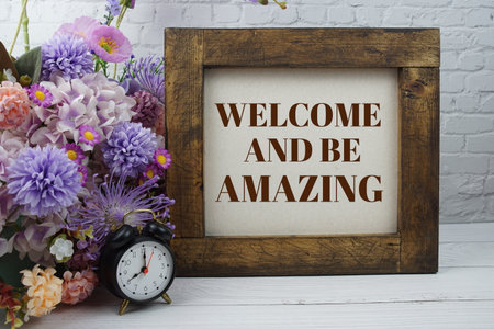 Welcome and be amazing text message motivational and inspiration quote with flowers decoration on white brick wall and wooden backgroundの写真素材