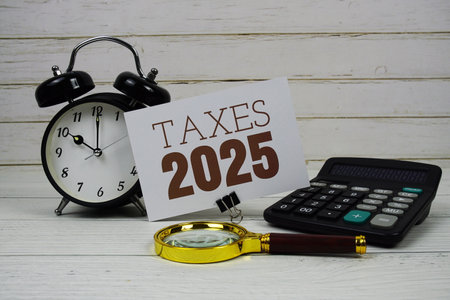 Taxes 2025 text on paper card with alarm clock, calculator and magnifying glass on wooden backgroundの写真素材
