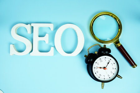 Seo letters (Search Engine Optimization) with magnifying glass and alarm clock on blue backgroundの写真素材