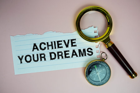 Achieve Your Dreams text written on paper with compass and magnifying glass flat lay on pink background