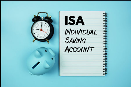 ISA abbreviation of Individual Saving Account text on notebook paper and piggy bank and alarm clock on blue backgroundの写真素材