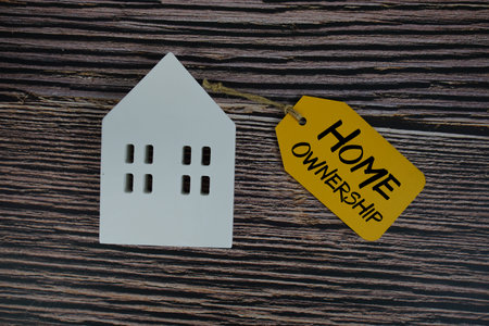Home Ownership text on price tag with white house model flat lay on wooden background