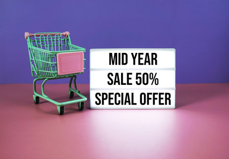 Mid Year Sale 50% off Special Offer text on LED lightbox with trolley shopping cart on pink and...