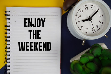 Enjoy The Weekend text written on notepad with alarm clock on blue backgroundの写真素材