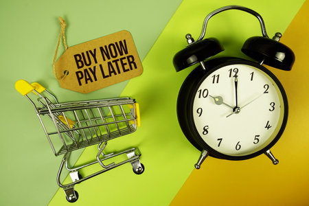 Buy Now Pay Later text on price tag with alarm clock and trolley shopping cart on green, yellow geometric backgroundの写真素材