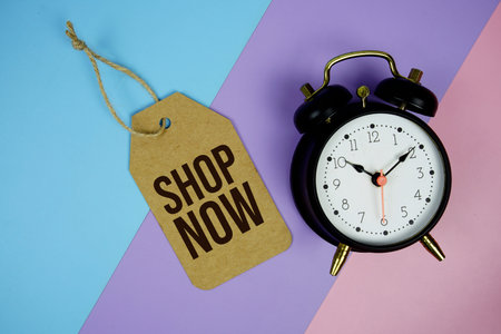 Shop Now text on price tag with alarm clock and trolley shopping cart on pink, purple, blue...