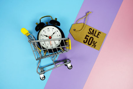 Sale 50% text on price tag with alarm clock and trolley shopping cart on pink, purple, blue...
