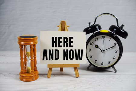 Here And Now text with hourglass and alarm clock on wooden backgroundの写真素材