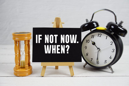If Not Now. When? text with hourglass and alarm clock on wooden backgroundの写真素材